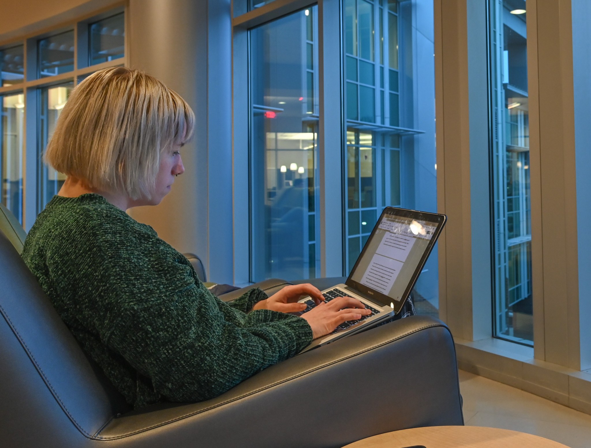 Blonde woman sitting in an easy chair in front of large windows with a laptop computer.